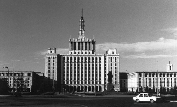 Black-and-white photograph of Casa Scânteii: a Stalinist tower block with a spire, flanked by lower wings, a statue on a plinth in front and a white car in the foreground.